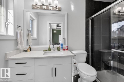 The bathroom features a vanity with a white countertop, a mirror with an overhead light fixture, and a shower with dark tiled walls and a glass enclosure - 1778 25A Street, Edmonton, AB - Indoor Photo Showing Bathroom