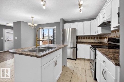 Kitchen with appliances with stainless steel finishes, dark countertops, a textured ceiling, white cabinetry, and a kitchen island with sink - 150 Acacia Circle, Leduc, AB - Indoor Photo Showing Kitchen With Double Sink