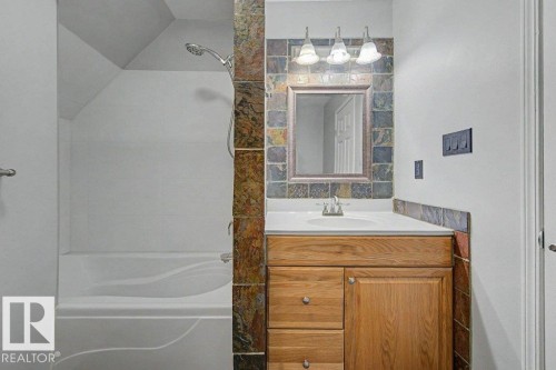Bathroom featuring a white bathtub with a showerhead, a vanity with a white countertop and wood-finish cabinetry, a framed mirror, and tile accents - Edmonton, AB - Indoor Photo Showing Bathroom