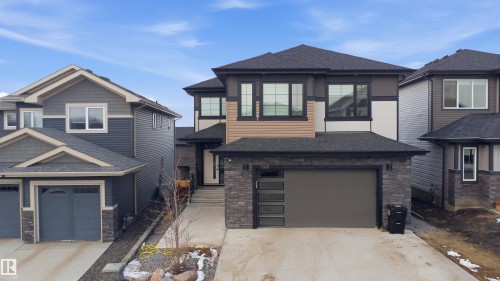 Contemporary two-story residence featuring a dark gray roofline, a mixed-material exterior with tan siding and stone veneer accents, and a dark gray garage door with frosted glass panels - 1210 Peregrine Terrace, Edmonton, AB - Outdoor With Facade