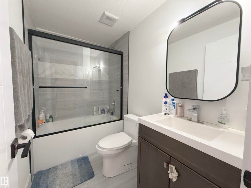 Modern bathroom featuring a dark wood-finish vanity with an integrated white sink, a contemporary black-framed mirror with integrated lighting, and a bathtub with a clear glass sliding enclosure - 1210 Peregrine Terrace, Edmonton, AB - Indoor Photo Showing Bathroom