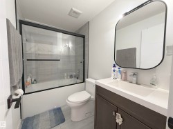 Modern bathroom featuring a dark wood-finish vanity with an integrated white sink, a contemporary black-framed mirror with integrated lighting, and a bathtub with a clear glass sliding enclosure - 