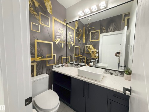 Powder room featuring a striking botanical-patterned accent wall, a wide vanity mirror with multi-bulb lighting, and a contemporary vessel sink - 1210 Peregrine Terrace, Edmonton, AB - Indoor Photo Showing Bathroom