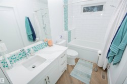 Bathroom featuring a white subway tile shower surround with an integrated accent tile stripe, a white vanity with an integrated sink, a large vanity mirror, and wood-finish flooring - 