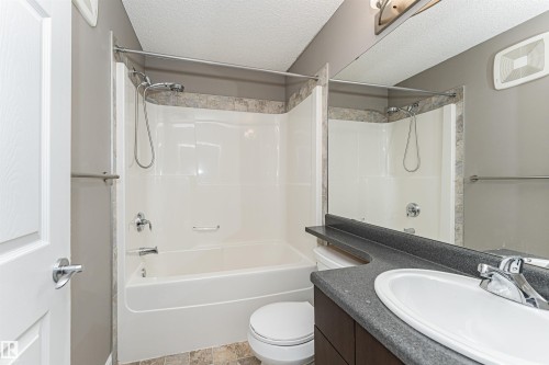 Bathroom featuring a white tub-shower combination with a textured tile surround, a dark wood-finish vanity with a dark countertop, an integrated sink, and chrome fixtures - 110 3625 144 Avenue, Edmonton, AB - Indoor Photo Showing Bathroom