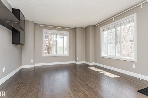 Spacious room featuring rich wood-finish flooring, two large windows with white trim, and a built-in wall unit - 110 3625 144 Avenue, Edmonton, AB - Indoor Photo Showing Other Room