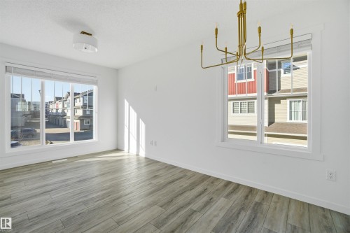 Spacious room featuring wood-finish flooring, two large windows with white trim, a modern brass chandelier, and a semi-flush mount ceiling light - 64 17832 78 Street, Edmonton, AB - Indoor Photo Showing Other Room