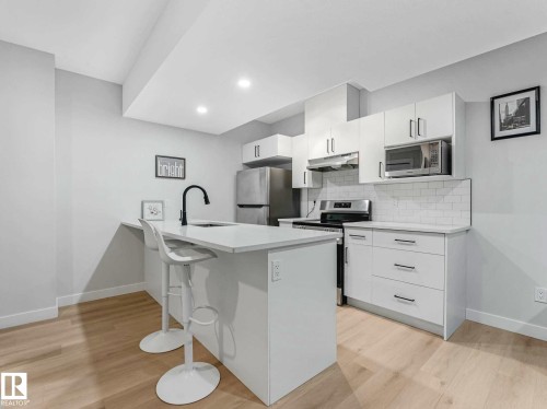 Kitchen featuring an island with a built-in sink and a matte black faucet, stainless steel appliances, white cabinetry with silver hardware, a subway tile backsplash, and light wood-finish flooring - 15731 109 Avenue, Edmonton, AB - Indoor Photo Showing Kitchen With Upgraded Kitchen