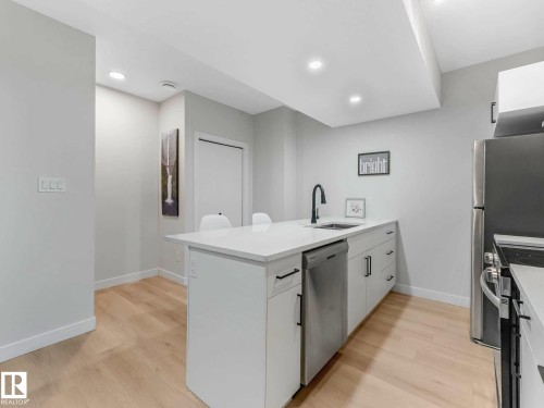 Kitchen island featuring a single-basin sink with a matte black faucet, white shaker-style cabinetry, stainless steel dishwasher, and light wood-finish flooring - 15731 109 Avenue, Edmonton, AB - Indoor Photo Showing Kitchen