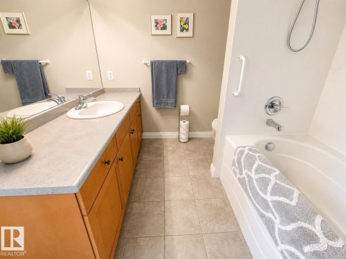 Bathroom featuring a combination tub/shower with chrome fixtures, a single vanity with an integrated sink, a light-toned countertop, and wood-finish cabinetry - 301 9803 96A Street, Edmonton, AB - Indoor Photo Showing Bathroom