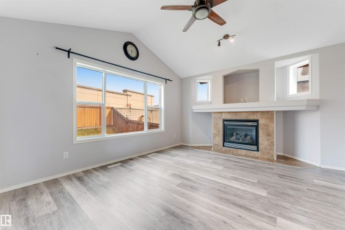 Bright living space featuring vaulted ceilings, a large picture window, wood-finish flooring, a ceiling fan, and a tiled fireplace surround with an integrated mantel and display niches - 123 Rue Masson, Beaumont, AB - Indoor Photo Showing Living Room With Fireplace