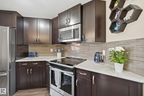 Kitchen featuring dark wood cabinetry, light-toned countertops, a glass tile backsplash, and stainless steel appliances - 481 Sheppard Boulevard, Leduc, AB - Indoor Photo Showing Kitchen With Upgraded Kitchen