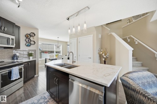 Kitchen featuring a central island with dual sinks and a stainless steel dishwasher, dark wood-finish cabinetry, stainless steel appliances, a mosaic tile backsplash, and wood-finish flooring - 481 Sheppard Boulevard, Leduc, AB - Indoor Photo Showing Kitchen With Double Sink With Upgraded Kitchen