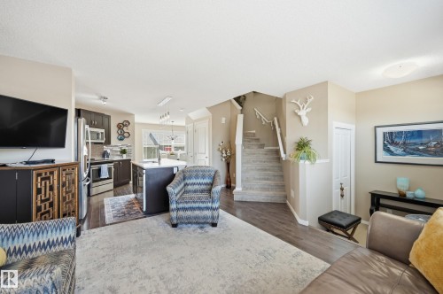 Open-concept living space featuring wood-finish flooring, a kitchen with dark cabinetry and stainless steel appliances, a central kitchen island with a sink, and a carpeted staircase with white railings - 481 Sheppard Boulevard, Leduc, AB - Indoor