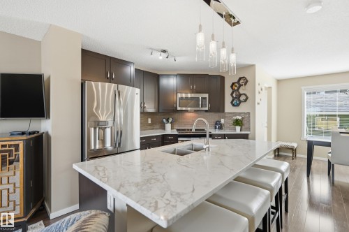 Kitchen featuring a large central island with a double basin sink, dark wood cabinetry, stainless steel appliances, a tiled backsplash, and wood-finish flooring - 481 Sheppard Boulevard, Leduc, AB - Indoor Photo Showing Kitchen With Double Sink With Upgraded Kitchen