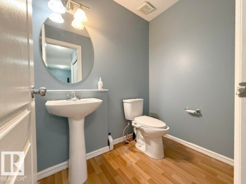 This property features a bathroom with light wood flooring, a white pedestal sink, a toilet, and a circular mirror - 3347 24 Avenue, Edmonton, AB - Indoor Photo Showing Bathroom