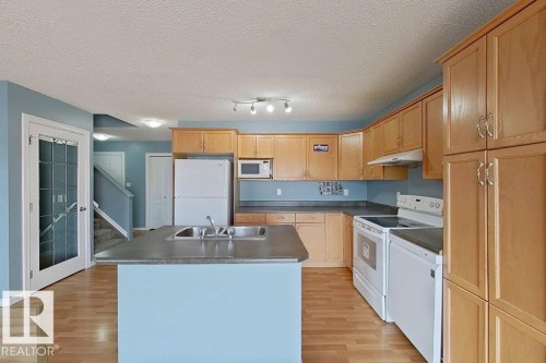 The kitchen features light wood cabinetry, a central island with a double sink, and white appliances including a refrigerator, oven, and microwave - 3347 24 Avenue, Edmonton, AB - Indoor Photo Showing Kitchen With Double Sink