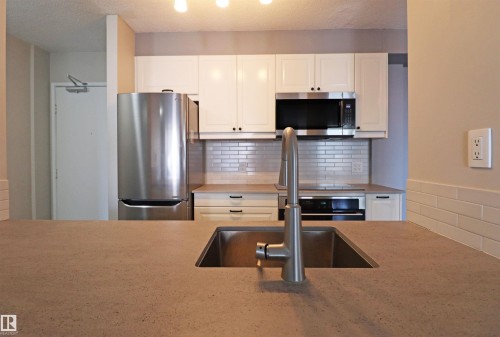 Kitchen featuring a large stainless steel sink with a modern faucet, light-toned countertop, white cabinetry with dark hardware, stainless steel appliances, and a subway tile backsplash - 901 10160 116 Street, Edmonton, AB - Indoor Photo Showing Kitchen