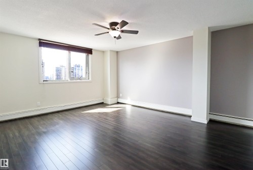 Spacious room featuring dark wood-finish flooring, a ceiling fan with integrated lighting, and a large window with a brown roller shade - 901 10160 116 Street, Edmonton, AB - Indoor Photo Showing Other Room