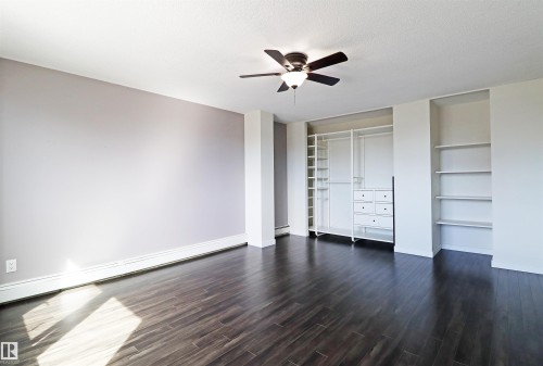 Spacious room featuring dark wood-finish flooring, a ceiling fan with integrated lighting, and a built-in closet organization system with shelving and drawers - 901 10160 116 Street, Edmonton, AB - Indoor