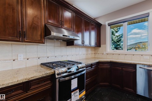 Dark wood-finish cabinetry with crown molding, stainless steel range with matching range hood, light-toned stone countertops, tiled backsplash with decorative border, and black tile flooring - 2408 Tegler Green, Edmonton, AB - Indoor Photo Showing Kitchen