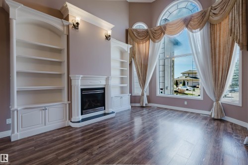 Two-story window with arched transom, custom drapery, wood-finish flooring, built-in shelving, and a fireplace with a white mantle - 2408 Tegler Green, Edmonton, AB - Indoor With Fireplace
