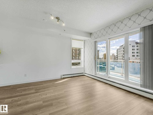 Bright living area featuring wood-finish flooring, white painted walls, a large window with a roller shade, and a three-panel glass sliding door leading to an exterior balcony - 305 10504 99 Avenue, Edmonton, AB - Indoor Photo Showing Other Room
