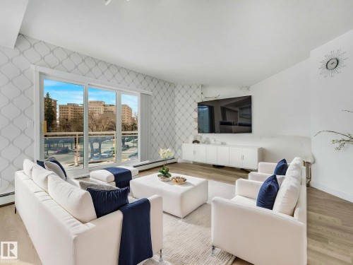 Living area featuring expansive sliding glass doors, patterned accent wall, light wood-finish flooring, a wall-mounted media console, and a contemporary wall clock - 305 10504 99 Avenue, Edmonton, AB - Indoor Photo Showing Living Room