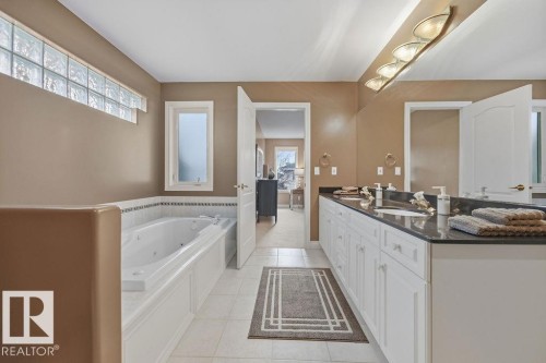 Bathroom featuring a jetted tub with tiled surround, a glass block window, a dual-sink vanity with dark countertop, white cabinetry, and white tile flooring - 11551 14 Avenue, Edmonton, AB - Indoor Photo Showing Bathroom