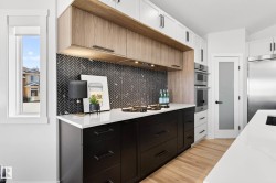 Modern kitchen featuring two-tone cabinetry with wood-finish upper cabinets and matte black lower cabinets, a herringbone tile backsplash, white countertops, and wood-finish flooring - 