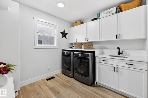 Dedicated laundry area featuring wood-finish flooring, white cabinetry with matte black hardware, a built-in sink with a matte black faucet, and a quartz countertop with a tiled backsplash - 324 Cutts Crest, Edmonton, AB - Indoor Photo Showing Laundry Room