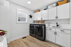 Dedicated laundry area featuring wood-finish flooring, white cabinetry with matte black hardware, a built-in sink with a matte black faucet, and a quartz countertop with a tiled backsplash - 