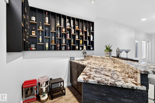 Built-in bar featuring a dark wood-finish base cabinet, a natural stone countertop, a stone veneer bar front, an integrated sink, and an illuminated display shelving system - 324 Cutts Crest, Edmonton, AB - Indoor