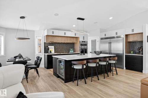 Spacious kitchen featuring a large island with seating, a built-in beverage cooler, stainless steel appliances, a herringbone tile backsplash, and light wood-finish flooring - 324 Cutts Crest, Edmonton, AB - Indoor