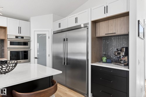 Gourmet kitchen featuring a large stainless steel refrigerator, double wall ovens, white cabinetry with black hardware, a black herringbone tile backsplash, and light wood-finish flooring - 324 Cutts Crest, Edmonton, AB - Indoor Photo Showing Kitchen With Upgraded Kitchen