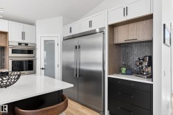 Gourmet kitchen featuring a large stainless steel refrigerator, double wall ovens, white cabinetry with black hardware, a black herringbone tile backsplash, and light wood-finish flooring - 