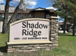 Stone veneer base with a stucco-finish sign, featuring mature trees and multi-story buildings with pitched roofs - 