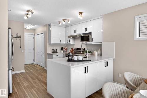 Modern kitchen featuring white high-gloss cabinetry with matte black hardware, a center island with an integrated sink and gooseneck faucet, light-toned countertops, and wood-finish flooring - 7723 7A Avenue, Edmonton, AB - Indoor Photo Showing Kitchen