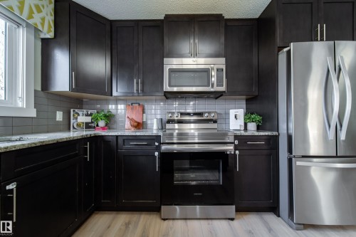 Kitchen featuring dark cabinetry, stainless steel appliances, speckled countertops, a tile backsplash, and light wood-finish flooring - 117 2229 44 Avenue, Edmonton, AB - Indoor Photo Showing Kitchen With Stainless Steel Kitchen With Upgraded Kitchen