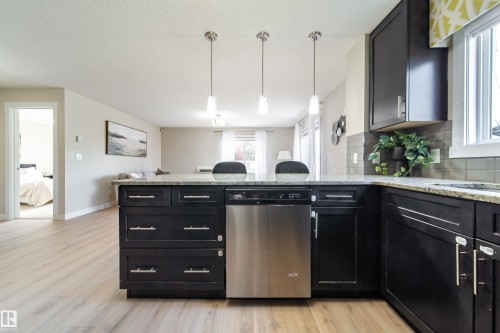 Kitchen island with a granite-style countertop, stainless steel dishwasher, dark wood-finish cabinetry, and three pendant lights - 117 2229 44 Avenue, Edmonton, AB - Indoor Photo Showing Kitchen With Upgraded Kitchen