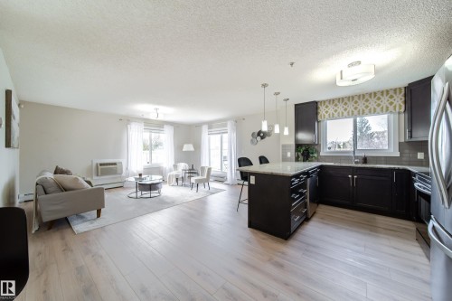 Open-concept living area featuring light wood-finish flooring, a kitchen island with pendant lighting, dark cabinetry, stainless steel appliances, and multiple windows - 117 2229 44 Avenue, Edmonton, AB - Indoor
