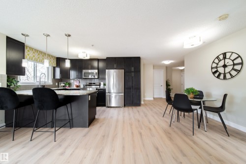 Open-concept kitchen and dining area featuring dark cabinetry, stainless steel appliances, a kitchen island with seating, pendant lighting, and wood-finish flooring - 117 2229 44 Avenue, Edmonton, AB - Indoor