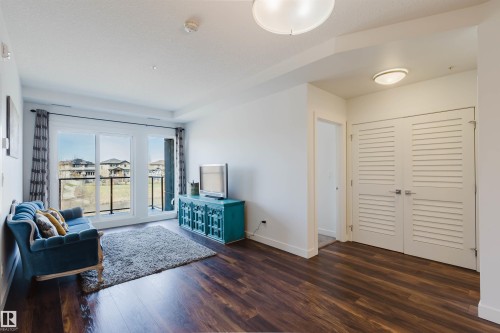 Living area featuring rich wood-finish flooring, white walls, and a large sliding glass door with a balcony and black metal railing - 205 1316 Windermere Way, Edmonton, AB - Indoor Photo Showing Other Room