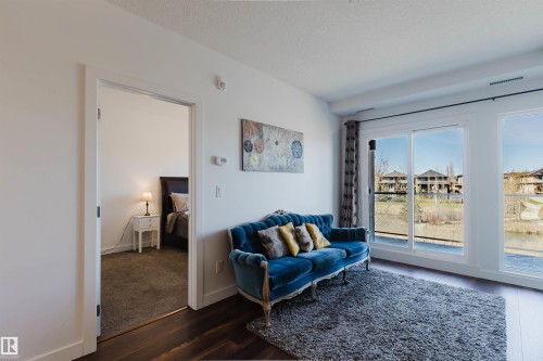 Expansive windows illuminate this living space, featuring dark wood-finish flooring, white walls, and a doorway to an adjoining carpeted room - 205 1316 Windermere Way, Edmonton, AB - Indoor