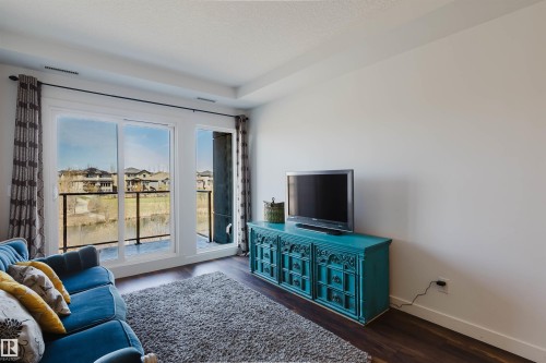 Spacious living area featuring expansive glass sliding doors leading to a balcony with glass railings, hardwood flooring, and a tray ceiling - 205 1316 Windermere Way, Edmonton, AB - Indoor
