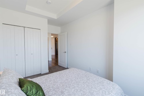 White-painted bedroom featuring a tray ceiling, bi-fold closet doors, and light gray carpeting - 205 1316 Windermere Way, Edmonton, AB - Indoor Photo Showing Bedroom