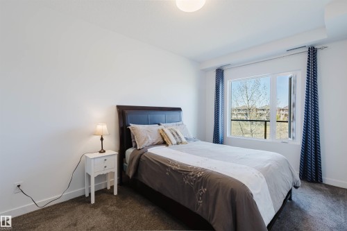 Bedroom featuring a large window, white walls, and dark carpeting - 205 1316 Windermere Way, Edmonton, AB - Indoor Photo Showing Bedroom