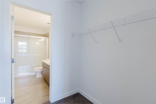 Walk-in closet with wire shelving and dark carpeting, opening to a bathroom featuring a shower with sliding glass doors, a vanity with under-sink cabinetry, and tile flooring - 205 1316 Windermere Way, Edmonton, AB - Indoor