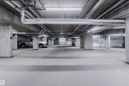 Spacious underground parking garage featuring concrete pillars and ceiling, bright overhead lighting, and visible utility conduits - 205 1316 Windermere Way, Edmonton, AB - Indoor Photo Showing Garage
