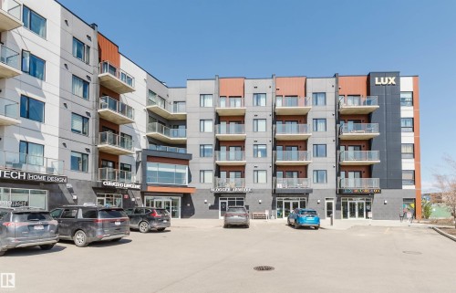 Contemporary multi-story building featuring a gray and white facade with architectural accents, multiple balconies with glass railings, ground-floor retail spaces, and an on-site parking area - 205 1316 Windermere Way, Edmonton, AB - Outdoor With Balcony With Facade